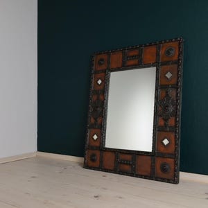 Pu&ograve; includere: Specchio decorativo quadrato con cornice marrone effetto pelle e decorazioni in metallo scuro. Lo specchio ha una superficie riflettente rettangolare ed &egrave; appoggiato a una parete verde scuro.