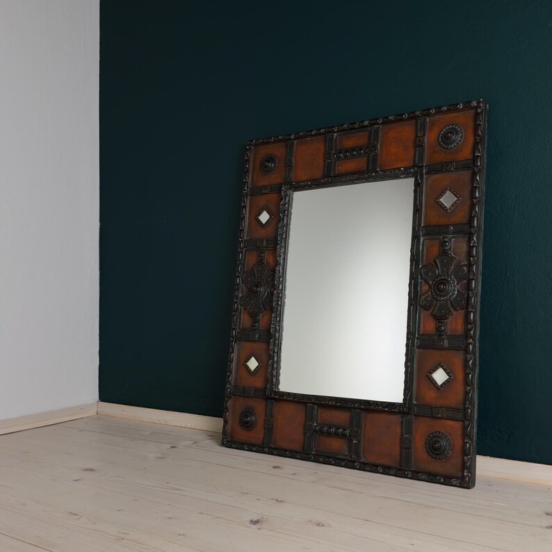Pu&ograve; includere: Specchio decorativo quadrato con cornice marrone effetto pelle e decorazioni in metallo scuro. Lo specchio ha una superficie riflettente rettangolare ed &egrave; appoggiato a una parete verde scuro.