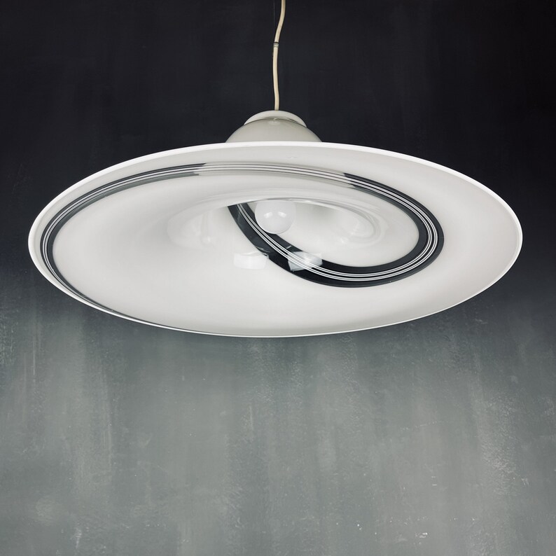 May include: A white ceiling light fitting with a swirling black and white design. The light fitting is suspended from the ceiling by a white cord.
