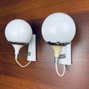 Pode incluir: Dois candeeiros de parede de globo branco com detalhes cromados e bases de cor creme. Os candeeiros s&atilde;o montados em placas retangulares brancas.