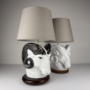 May include: Two ceramic table lamps. Each lamp features a ram's head base with a white finish and black accents. The lampshades are a light beige color. The bases are set on a dark brown, oval-shaped base.