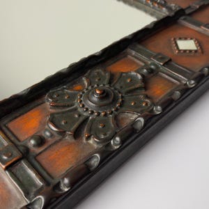 Pu&ograve; includere: Primo piano di una cornice decorativa per specchio con un motivo floreale ornato marrone scuro. La cornice ha una forma rettangolare con una finitura marrone-rossastra invecchiata e un bordo nero. La superficie dello specchio &egrave; visibile.