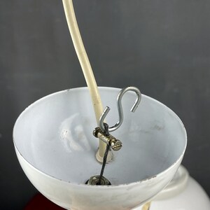 May include: Close-up of a white lampshade with a metal hook and a white cord attached to it. The cord is threaded through a metal clamp.
