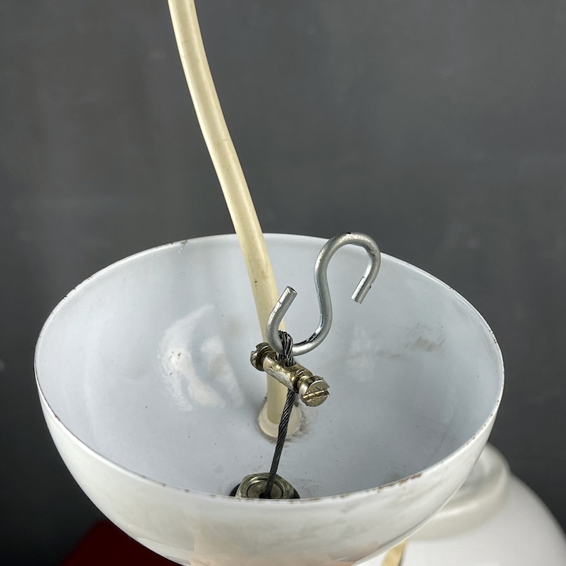 May include: Close-up of a white lampshade with a metal hook and a white cord attached to it. The cord is threaded through a metal clamp.