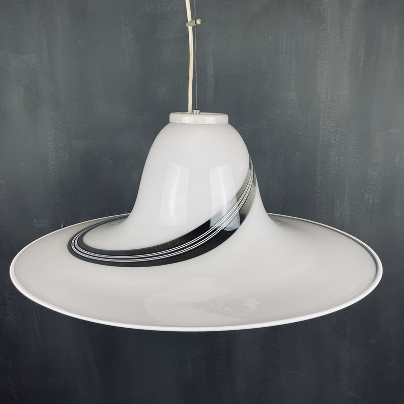 May include: A white pendant light with a wide, flat, saucer-shaped shade. The shade has a black and white striped design.