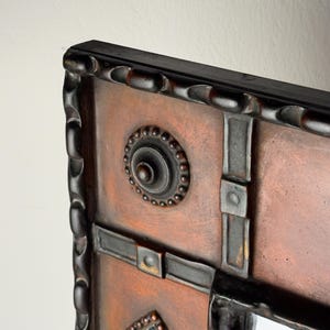 Pu&ograve; includere: Primo piano di uno specchio decorativo con cornice in legno marrone scuro. La superficie dello specchio ha una finitura color rame con accenti marrone scuro e un design circolare e ornato al centro. La cornice ha un motivo strutturato e intagliato.