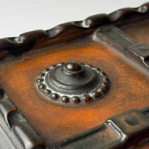 Pu&ograve; includere: Primo piano di un cofanetto decorativo in stile antico. La scatola presenta un bordo ornato marrone scuro e un ornamento metallico centrale circolare con un centro rialzato e un bordo perlato. La scatola ha un colore marrone rossastro caldo.