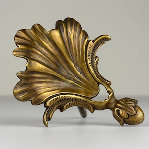 May include: A detailed, antique brass-colored decorative object. The piece features a large, fan-shaped element with intricate veining, connected to a smaller, rounded form. The design has a vintage aesthetic, with a focus on ornate details and a warm, metallic finish.