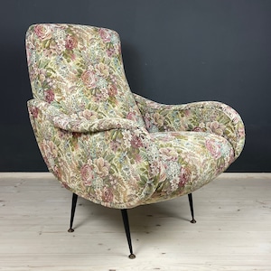Op de afbeelding: Een fauteuil met een bloemenpatroon en zwarte metalen poten. De stoel heeft een gebogen rugleuning en een brede zitting.