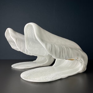 May include: A white ceramic sculpture of two abstract shapes resembling waves or shells. The sculpture is smooth and has a glossy finish.