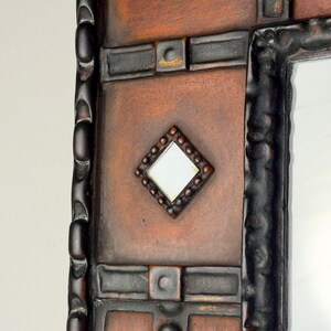 Pu&ograve; includere: Primo piano di uno specchio decorativo con una cornice rustica e strutturata. La cornice presenta tonalit&agrave; marrone scuro e rame, con un piccolo specchio a forma di diamante al centro. Il design include forme geometriche e un'estetica vintage.