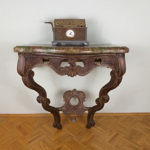 Op de afbeelding: Een bruine houten consoletafel met sierlijke houtsnijwerk en een marmeren blad. De tafel heeft een gebogen ontwerp en staat op vier poten. Een klein antiek klokje staat op de tafel.