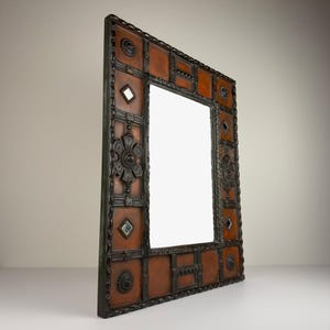 Pu&ograve; includere: Uno specchio rettangolare decorativo con una cornice marrone simile alla pelle. La cornice &egrave; impreziosita da decorazioni in metallo scuro, tra cui forme geometriche, motivi floreali e piccoli specchi quadrati. Lo specchio &egrave; angolato, mostrando la sua superficie riflettente.