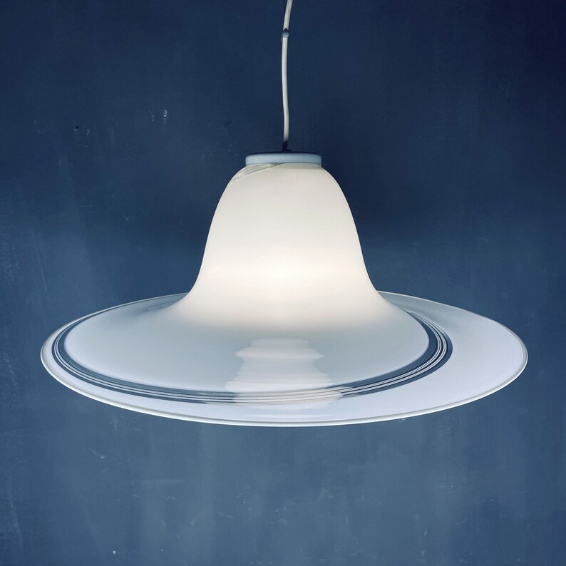 May include: A white glass pendant light with a wide, circular, saucer-shaped base and a bell-shaped top. The base has a thin, silver-coloured band around the edge.