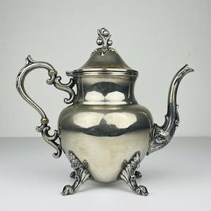 Peut inclure: Une théière en argent avec un design orné et trois pieds. La théière a un couvercle avec un motif floral sur le dessus.