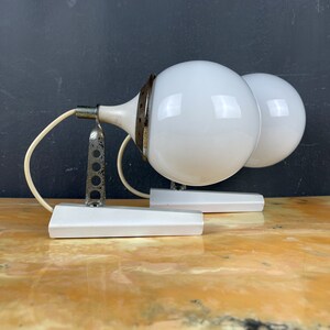 Pode incluir: Dois candeeiros de parede brancos com c&uacute;pulas redondas de vidro branco. Os candeeiros t&ecirc;m uma base de metal prateado e uma placa de montagem de pl&aacute;stico branco.