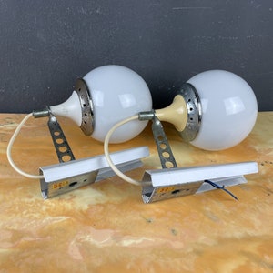 Pode incluir: Um par de candeeiros de parede brancos em forma de globo com detalhes prateados e creme. Os candeeiros t&ecirc;m um design &uacute;nico com uma base circular e um bra&ccedil;o curvo. Os candeeiros est&atilde;o montados numa superf&iacute;cie branca.