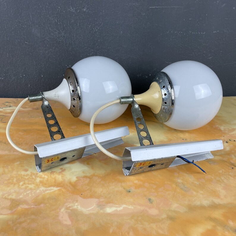 Pode incluir: Um par de candeeiros de parede brancos em forma de globo com detalhes prateados e creme. Os candeeiros t&ecirc;m um design &uacute;nico com uma base circular e um bra&ccedil;o curvo. Os candeeiros est&atilde;o montados numa superf&iacute;cie branca.