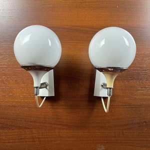Pode incluir: Dois candeeiros de parede de globo branco com uma base de metal prateado e um cabo branco. Os candeeiros est&atilde;o montados numa superf&iacute;cie de madeira.