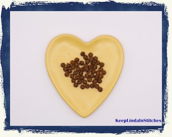 Puede incluir: Un plato amarillo en forma de corazón lleno de pequeñas campanas marrones. El plato está sobre una superficie blanca, con un borde azul. El texto "KeepLindaInStitches" es visible en la esquina inferior derecha.