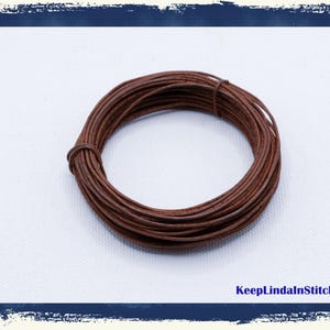 May include: A coil of brown leather cord, tightly wound, resting on a white surface. The cord appears to be thin and flexible, suitable for crafting or jewelry making. The image has a blue border and the text "KeepLindaInStitches".