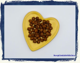 Puede incluir: Un plato amarillo en forma de corazón lleno de pequeñas campanas marrones. Las campanas están agrupadas. El plato está sobre un fondo blanco con un borde azul. El texto "KeepLindaInStitches" está en la esquina inferior derecha.