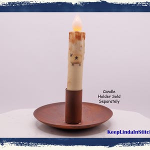 May include: A battery-operated candle with a flickering flame sits on a copper-colored plate. The candle has a cream-colored body with star and decorative accents. The text "Candle Holder Sold Separately" is visible.