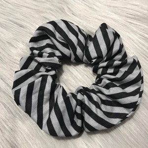 May include: A black and gray striped scrunchie. The fabric is gathered and elasticized, creating a circular shape. The stripes are evenly spaced and run diagonally across the fabric. This hair accessory is a stylish addition to any hairstyle.
