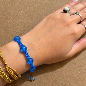 May include: A bright blue macrame bracelet featuring four round frosted beads, worn alongside two mustard yellow braided cord bracelets with metallic accents. The blue bracelet has adjustable pull strings finished with small silver-toned metal beads.