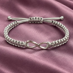 Könnte beinhalten: Ein hellgraues, geflochtenes Armband mit einem silberfarbenen Unendlichkeitssymbol in der Mitte. Das Armband verfügt über einen Schiebeknotenverschluss mit zwei verstellbaren Kordelenden, die jeweils mit einer kleinen silberfarbenen Perle versehen sind.