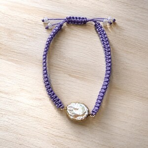 May include: A purple woven cord bracelet featuring a single oval-shaped iridescent white pearl set in a gold-toned metal frame. The bracelet includes an adjustable sliding knot closure with small clear beads at the ends of the pull cords.