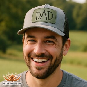 Könnte beinhalten: Grau-schwarze Trucker-Cap mit einem grünen Aufnäher mit der Aufschrift "DAD". Die Kappe hat einen gebogenen Schirm und eine Mesh-Rückseite. Die Kappe wird von einer lächelnden Person getragen.