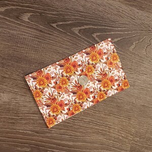 May include: A white fabric clutch with a floral pattern featuring orange, yellow, and brown flowers. The clutch has a coin on top of it.