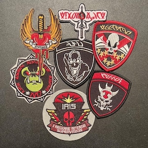 May include: A collection of nine embroidered patches featuring skulls, swords, and other graphic designs. The patches are mostly black, red, and white with some gold accents. The patches have various text including "VIXEN", "IRIS", "I REQUIRE INTENSE SUPERVISION", and "GAMMA".