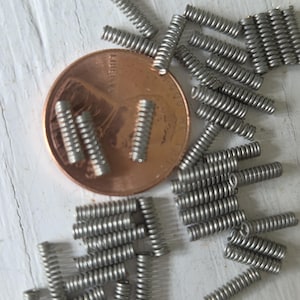May include: Close-up of numerous small, silver-colored coil springs scattered around a copper penny. The springs are tightly wound and appear to be made of metal. The penny is a US one-cent coin, showing the profile of Abraham Lincoln.