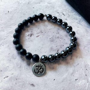 Może przedstawiać: Bransoletka z czarnych i srebrnych koralików z zawieszką w kształcie symbolu Om. Bransoletka wykonana jest z czarnych matowych koralików i srebrnych koralików hematytowych.