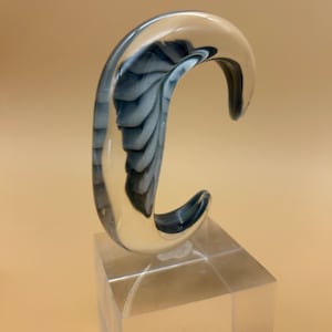 May include: A clear, curved glass sculpture with a swirling blue and white interior design. The sculpture is mounted on a clear, square acrylic base, creating a modern and artistic display piece.