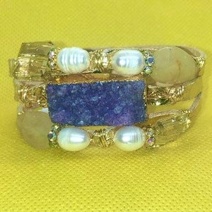 May include: A multi-strand wrap bracelet featuring a large, rectangular, deep purple stone, creamy white pearls, and yellow-toned stones. The bracelet has gold-colored metal accents and is set against a yellow background.