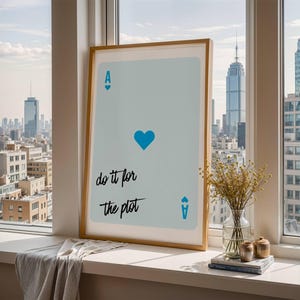 May include: A framed art print featuring a light blue playing card design with a blue heart and the text "do it for the plot." The print is displayed on a windowsill with a city skyline in the background. A vase of dried flowers and books are also on the windowsill.