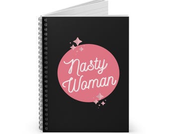 May include: A spiral-bound notebook with a black cover. The cover features a pink circle with the words "Nasty Woman" in white script. Pink starbursts and sparkles surround the circle.