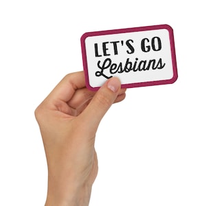 以下が含まれることがあります： ピンクの縁取りのある白いパッチで、黒い刺繍で「LET'S GO Lesbians」と書いてあります。