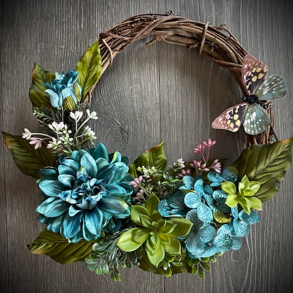 This small 10” grapevine wreath is the perfect touch for spring or summer.  Teal dahlia,  hydrangea, succulent accents & butterfly.