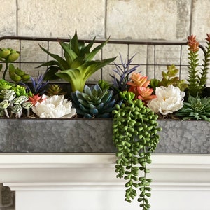 Puede incluir: Una maceta de metal galvanizado con una rejilla de alambre en la parte superior llena de una variedad de suculentas y flores artificiales, incluyendo flores blancas, rosas y naranjas.