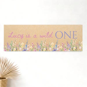 Könnte beinhalten: Ein rechteckiges Schild mit hellbraunem Kraftpapier-Hintergrund und dem Text Lucy is a wild ONE in violetter Schreib- und Blockschrift. Der untere Rand ist mit einer Aquarellillustration von bunten Wildblumen verziert.