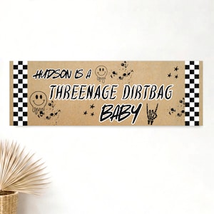 May include: A rectangular banner with a tan background and black and white checkered borders on the sides. The text reads "HUDSON IS A THREENAGE DIRTBAG BABY" in black, accompanied by black graphics of melting smiley faces, stars, and a skeleton hand.