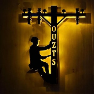 May include: A black metal silhouette of an electrician climbing a utility pole, with the word "OUTZS" vertically displayed. The pole and the top of the design are illuminated with a warm yellow light, creating a striking contrast against the dark background.