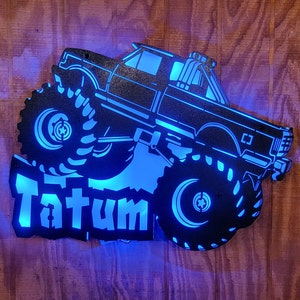 May include: A black metal silhouette of a monster truck with large tires and the name "Tatum" in white lettering. The truck is illuminated with blue LED lights.
