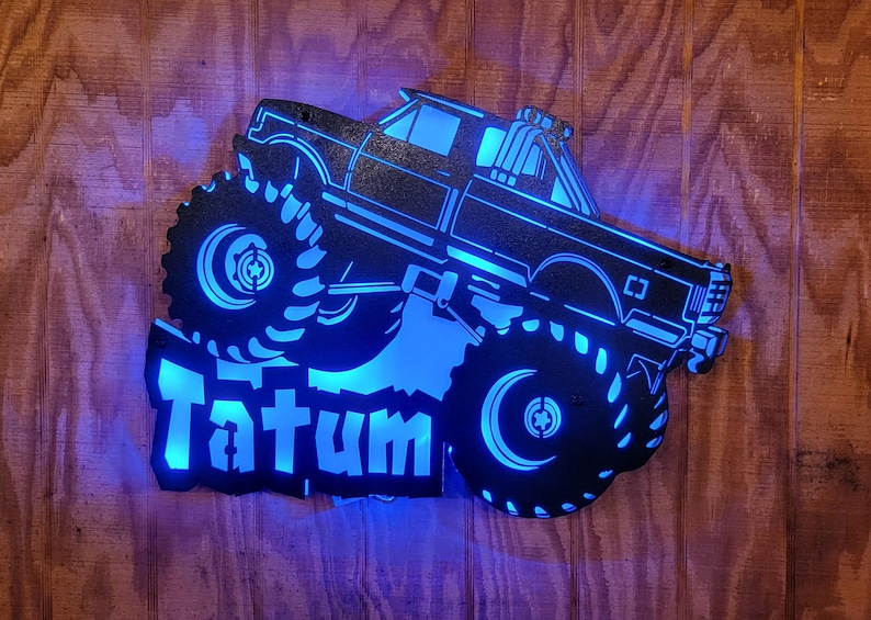May include: A black metal silhouette of a monster truck with large tires and the name "Tatum" in white lettering. The truck is illuminated with blue LED lights.