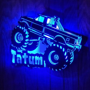 May include: A blue illuminated wooden cutout of a monster truck with the name Tatum in a stylized font.