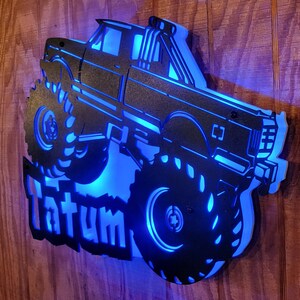 May include: A black metal silhouette of a monster truck with large tires and a blue light behind it. The name "Tatum" is written below the truck.